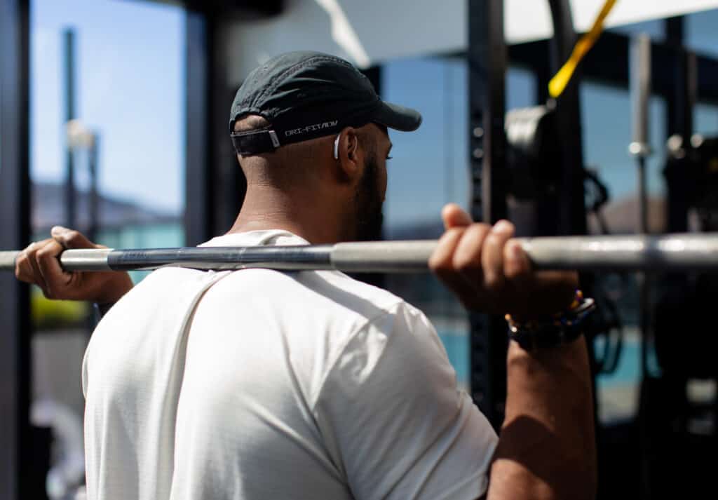 A man in a baseball cap is seen from behind, holding a baseball bat across his shoulders. He is wearing a slim silver hearing aid.