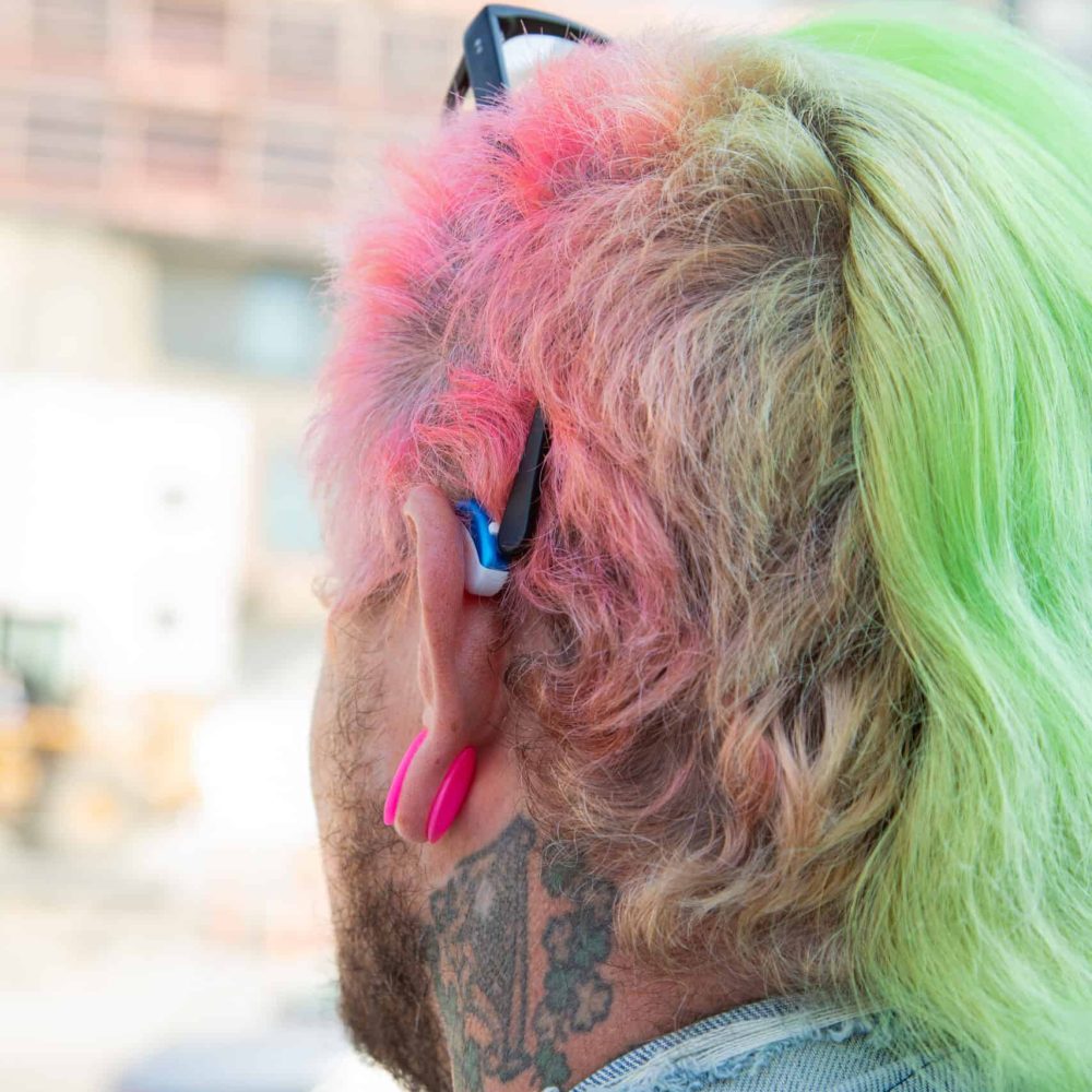 Man with green and pink hair wearing a blue hearing aid.